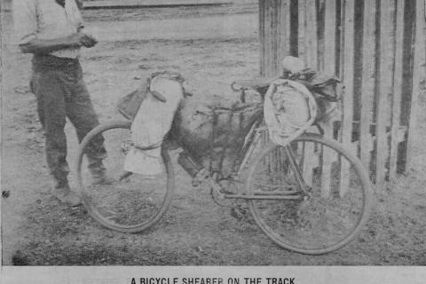 A C19th shearer stand next to his bicycle which is covered in bags and swags. 