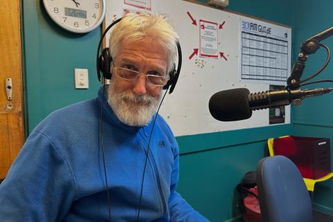 A man with white hari and a beard sits in front of a microphone. He is wearing headphones and looking at the camera