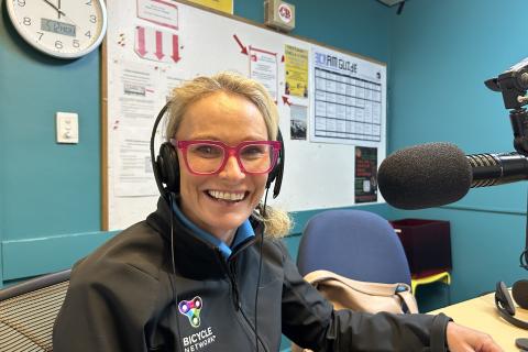 A woman with blonde hair and pink glasses faces the camera smiling. She is sitting in front of a microphone and wearing a black jackets with 'Bicycle Network' on it. 