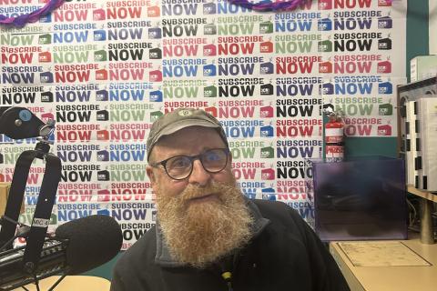 A bearded man sits at the desk with a microphone to one side of him. He is smiling and behind him the wall is covered in Subscribathon material. 