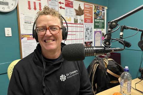 A smiling man wearing headphones sits next to a microphone in the 3CR studio. he is wearing a black sweatshirt with "Dr Cranky's Bikes for kids" written on it. 