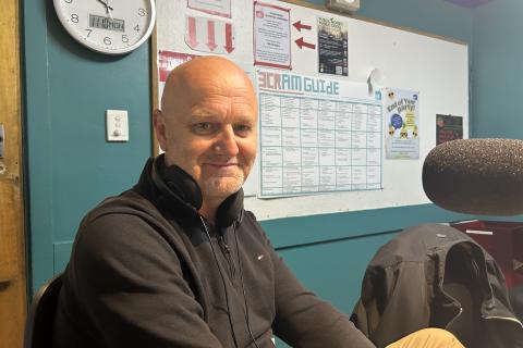 A man wearing a black top sits facing the camera and smiling
