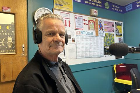 A man with grey hair is wearing headphones, seated in front of a microphone and turned towards the camera. 