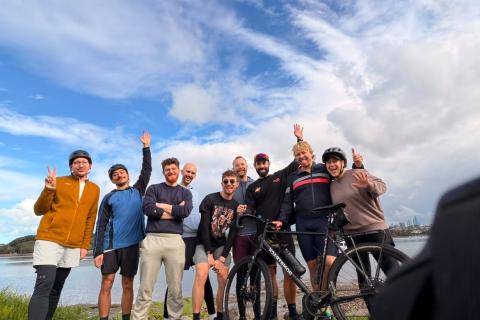 Group of casually attired bicycle riders smiles at the camera, clouds and blue sky behind them.