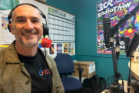 A man wearuing a t-shirt with a logo that says Good Cycles, is wearing headphones and sitting in the 3CR studio. He is smiling at the camera. 