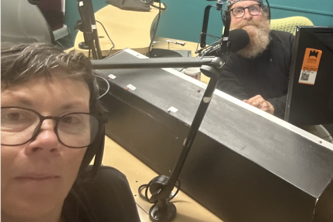 A woman wearing glasses looks into the camera, nehind her is a panelling dek and behind that a bearded man looking at the camera. 