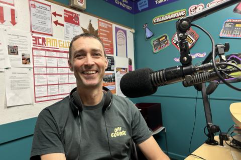 A smiling man in a green t-shirt with the logo "Off Course" on it sits next to a microphone and looks at the camera. 