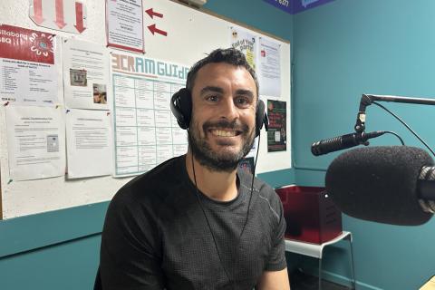 A man in a black t-shirt sits smailing at the camera. he has headphones on his head and is sitting in front of a microphone