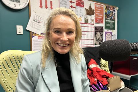 A smiling woman with blonde hair in a pale blue blazer sits in the studio at 3CR looking at the camera. 