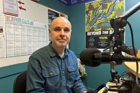A man sitting in front of a microphone faces the camera 