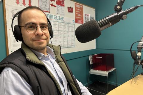 A man wearing headphones sits in front of a microphone and faces the camera