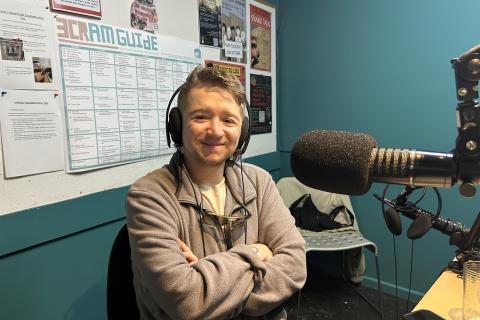 A young person with a beard sits in front of the microphone in the 3CR studio. Their arms are folded and they are smiling. 