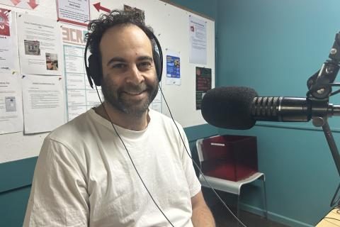 A bearded man in a white t-shirt siots in the 3CR studio. He has a microphone in front of him and is smiling at the camera. 