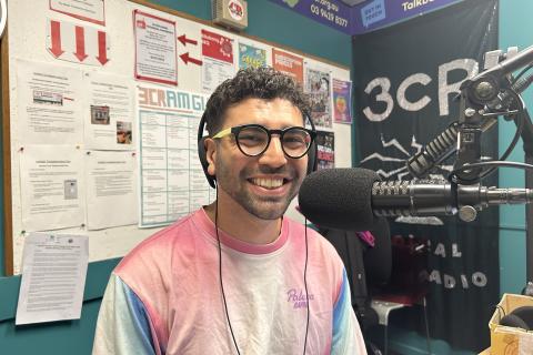 A smiling man wearing glasses and a dope longsleeved tee sits in front of a microphone in the studio at 3CR