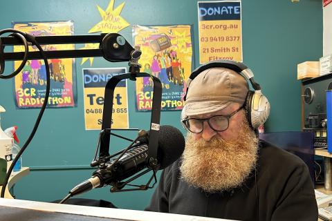 a bearded man wearing a cap sits at a microphone