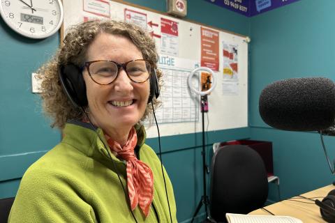 A smiling woman with curly hair sits in front of a microphone. She is wearing glasses, a green top and headphones. 