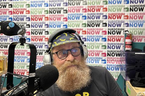 A bearded man wearing headphones sits in front of a microphone. behind him is wall poster with Subscribe on it repeatedly. 