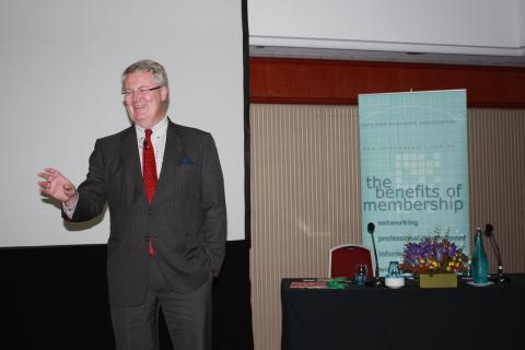 Man in a black suit and red tie smiling and standing at a conference 