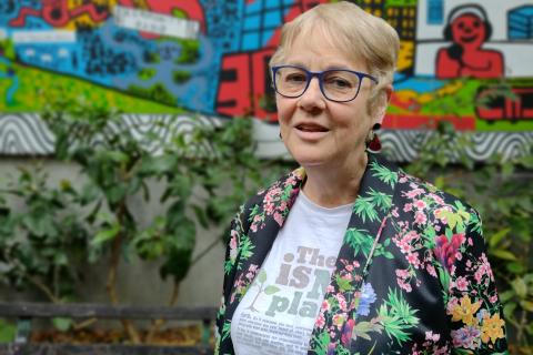An older woman wearing a colourful jacket and short hair looks at the camera, she's standing outside in front of a mural and she looks content