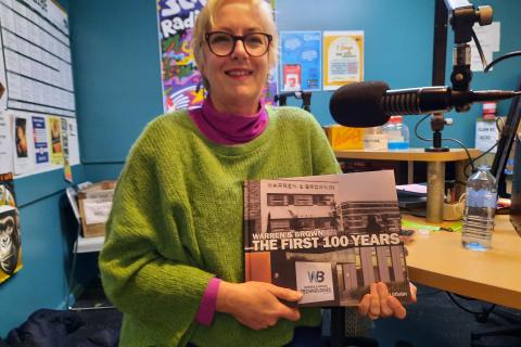 Deborah sits in studio 1 at 3CR holding her book and smiling at the viewer. She wears a green and pink jumper and sports black glasses