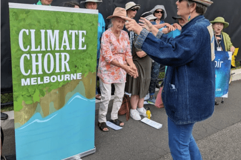 A photo of the Climate Choir performing. There is a big sign that says Climate Choir, a woman is conducting the choir and a group of people are singing. 