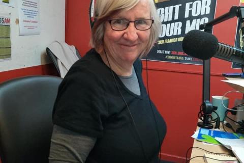 A woman with grey hair is looking at the camera, she is in a radio studio wearing headphones, glasses and a black shirt with a grey undershirt. 