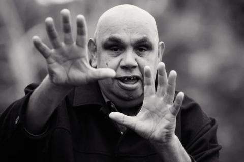 A black-and-white image of Kutcha with his arms raised in front of him making an expressive hand gesture towards the viewer