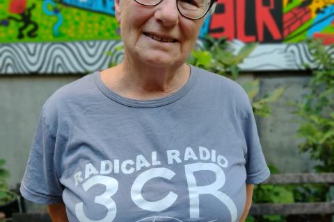 Linda Marks has short grey hair and glasses, she's pictured in the 3CR courtyard with a colourful mural behind her and is wearing a grey 3CR Radical Radio shirt