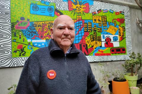 Percy stands in front of 3CR's new colourful mural in the courtyard. He wears a blue knitted jumper and sports a red YES badge