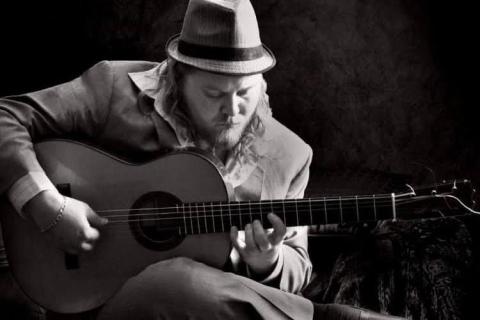 A black-and-white image of man playing the guitar. He sits down looking at the guitar with his right ankle crossed over his left knee. He wears a suit and sports a hat.