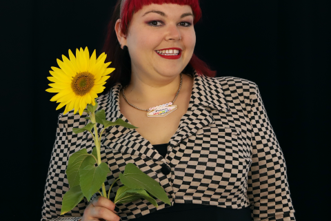 A woman stands straight on and looks into the camera holding a large sunflower up to her shoulder