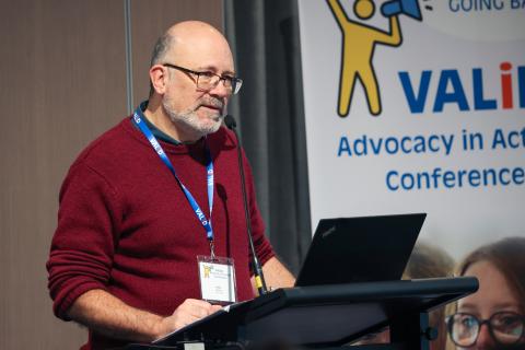 VALID CEO Fionn Skiotis, is standing at a podium and speaking into a microphone. He is wearing a red umper blue lanyard, and qlasses. Behind him is a white banner with vellow and blue graphics, text reads: 'VALID Advocacy in Action Conference'