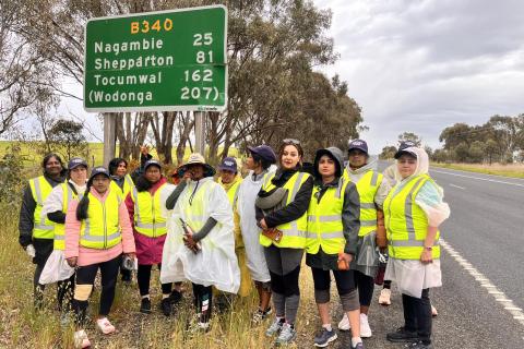 A photo of Refugee Women Action for Visa Equality wearing high vis vests, standing in front of a large road sign. They are standing on the side of a highway.
