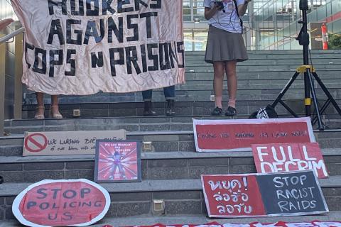 The foreground, steps in front of a corporate office building covered in handmade painted cardboard signs in various languages, two in English read: Stop policing us! and Full Decrim! The background shows two people holding a large homemade banner with black gaffer tape letters on light pink silky material that reads HOOKERS AGAINST COPS + PRISONS, with another person holding a microphone next to a speaker. The microphone has a little label on it that reads 3CR.