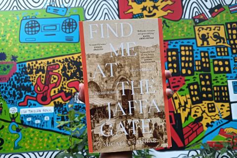 A copy of the book 'Find Me at the Jaffa Gate' by Micaela Sahhar, held in front of a colourful mural in the 3CR station courtyard.