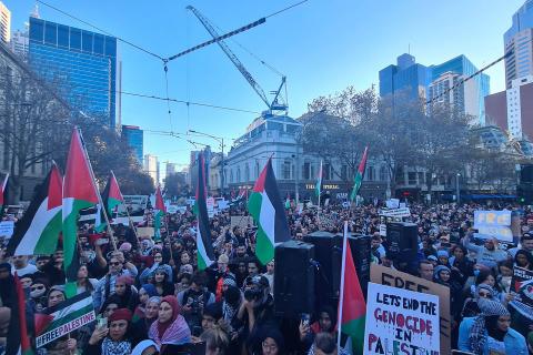 A photograph of the Spring Street and Bourke Street intersection in Narrm/Melbourne filled with people attending the Nakba Rally. Many are holding Palestinian flags and signs of solidarity with Palestine.