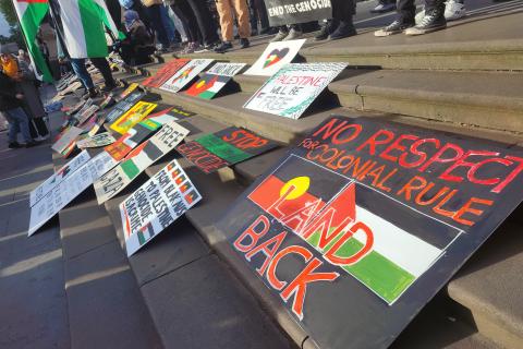 Painted signs on the steps of Parliament House with messages of solidarity between Aboriginal people and Palestine. The closest sign has the Aboriginal and Palestinian flags painted on it and says "No respect for colonial rule land back".