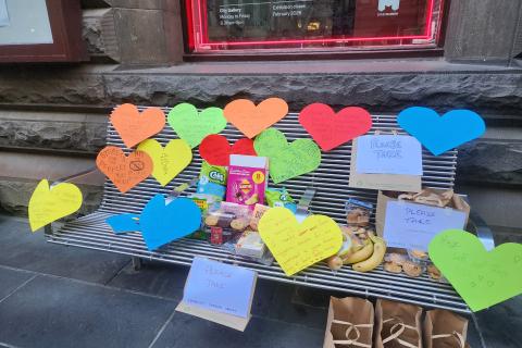 A city bench with large colourful paper hearts stuck all over it sharing messages of solidarity with Melbourne's homeless community. There is also an assortment of food on the bench.