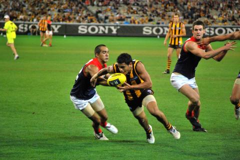 Hawthorn Indigenous AFL player Cyril Rioli pushes away a Melbourne player in the midst of an AFL match. He has the football tucked under his other arm and is about to take off running.