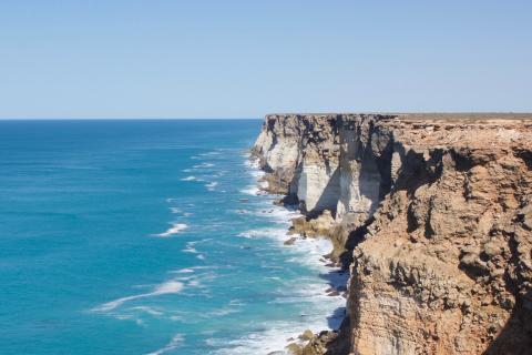 A photograph of the Great Australian Bight. To the right are steep rocky cliffs topped by a flat plain, and to the left is open water. The ocean is deep blue, growing darker towards the horizon. The sky is light blue and cloudless. Waves crash against the cliff face.