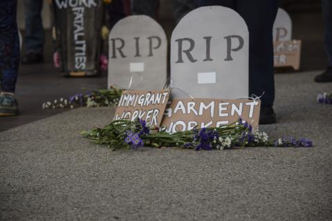 A photo of two cardboard tombstones with RIP written on them. A cardboard sign leans against the base of each tombstone, one reads "immigrant worker" and the other reads "garment worker". Flowers are laid below the signs.
