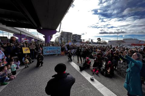 In the centre of the image we can see the back of Gaetano Greco as he addresses a gathering of people at the 2023 Hands Around Preston community protest to save the Preston Market from demolition and development. The gathering of people hold signs saying, 'Community Market,' and 'Honk to save our Preston Market'. 