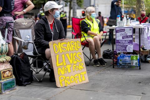 A photograph of a person sitting in a camping chair at a rally holding a sign which reads "DISABLD LIVES NOT FOR PROFIT". They are wearing a cap and a face mask and looking away from the camera. There is another masked person sitting behind and to the right of them, behind a table with water bottles, sanitiser and information.