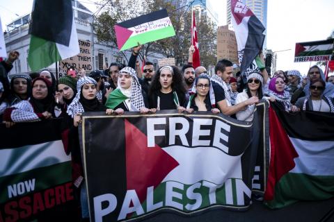 A photograph of a large crowd of protesters at a Free Palestine rally. Many are waving Palestinian flags and signs and wearing kuffiyehs. The front row of protesters are holding a large banner printed with the Palestinian flag overlaid with the text "Free Palestine".