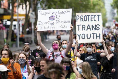 A photograph of a crowd at the Rainbow Rebellion in Melbourne to oppose the Religious Discrimination Bill. Two prominent signs are visible in the photo- one reads "respect my existence or expect our resistance, protect queer kids" and the other reads "protect trans youth".