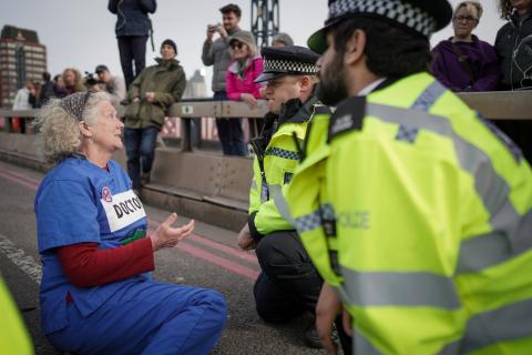 A photograph of a doctor in scrubs sitting on the ground on a bridge in the UK being confronted by two police officers in high-visibility jackets.