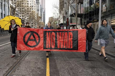 A long red banner reading 'NO WAR' in black text is held up by marching protesters. 