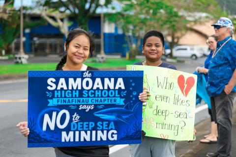 Two attendees stand with signs from a united gathering as a Stand with The Moana, held by Finafinau on 16th July, to call for the end of deep sea mining in waters off American Samoa. One sign reads 'The Samoana High School Finafinau Chapter says no to deep sea mining'. The second sign reads, 'Who even likes deep-sea mining?'