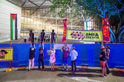 A photograph of protestors peacefully demonstrating outside the Brisbane Convention Centre where Land Forces is being held. Protestors are prevented from getting closer by a tall blue barricade, but they hold up black fibreglass casts of the figures of children and a colourful banner that reads 'Arms Dealers Export Terror!' A Palestinian flag hangs vertically to the left of the protestors.