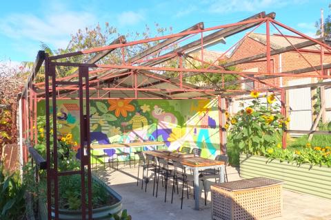 A photograph of an outdoor seating area at Oakhill Food Justice Farm bordered on either side by raised planter beds and with a large wall painted with a colourful mural. A metal roof frame sits over the space, but it is open to the elements. There are tables and chairs inside the space.